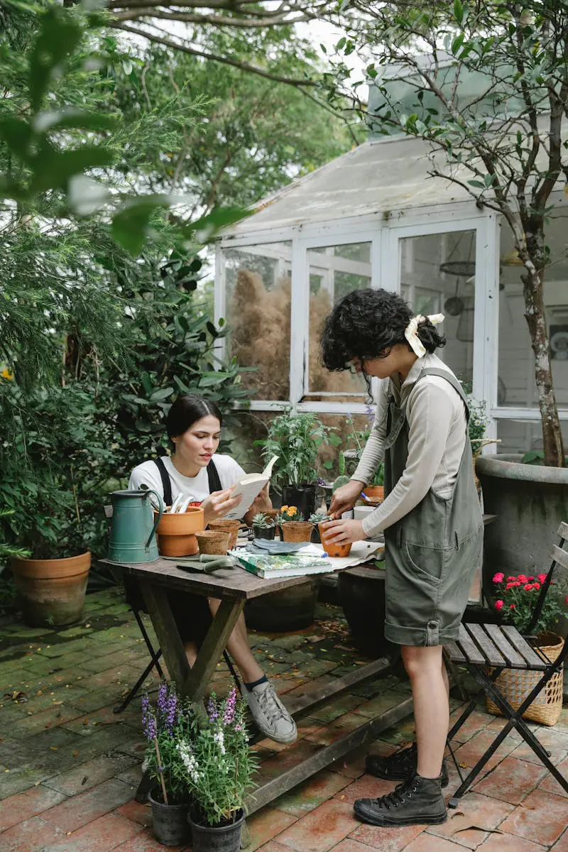 Full body of female workers cultivating sprouts in pots and reading gardening book at wooden table in backyard