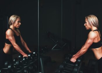 woman wearing black top top holding black dumbbells standing in front of mirror