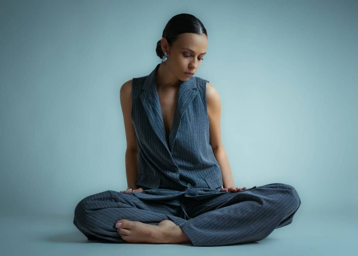 A woman in a vest and pants sits cross-legged.